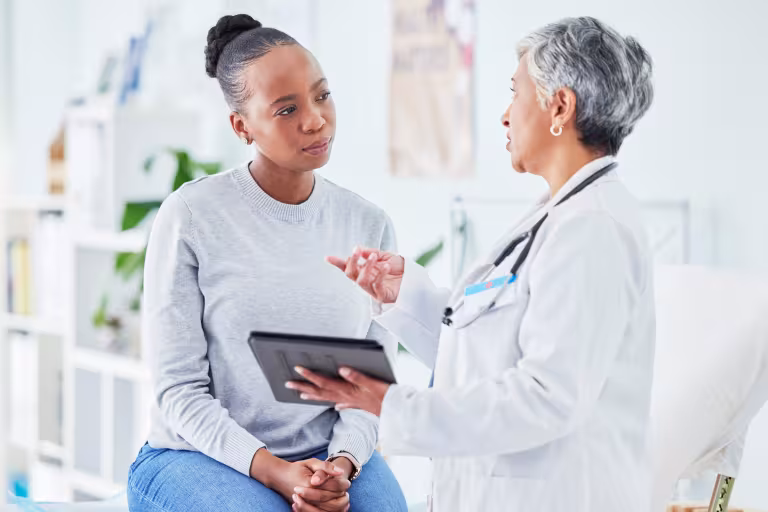A woman sits on an exam table listening to a doctor who is speaking to her and holding a tablet in her hand. 