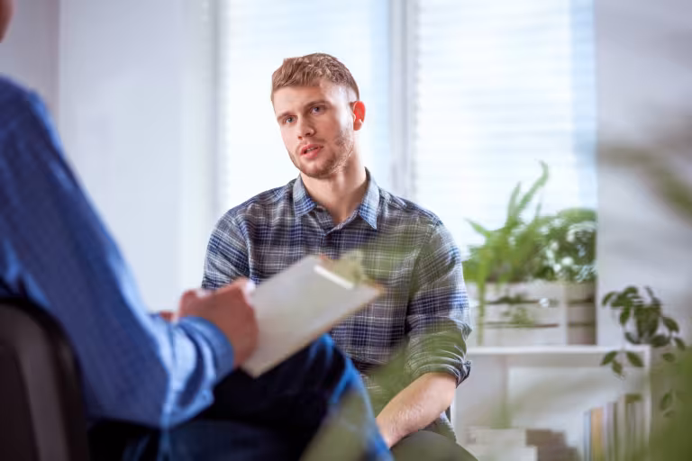 A young adult male sits in a therapist office, the therapist is out of frame but you can see their arm and a notepad for taking notes while the patient speaks.