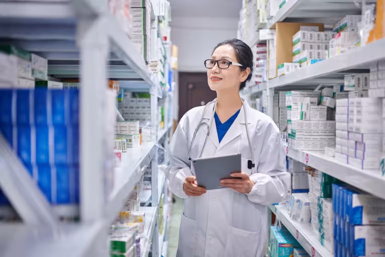 A female pharmacist walks down the aisles of a stocked pharmacy with a clipboard in hand while she looks for a prescription.