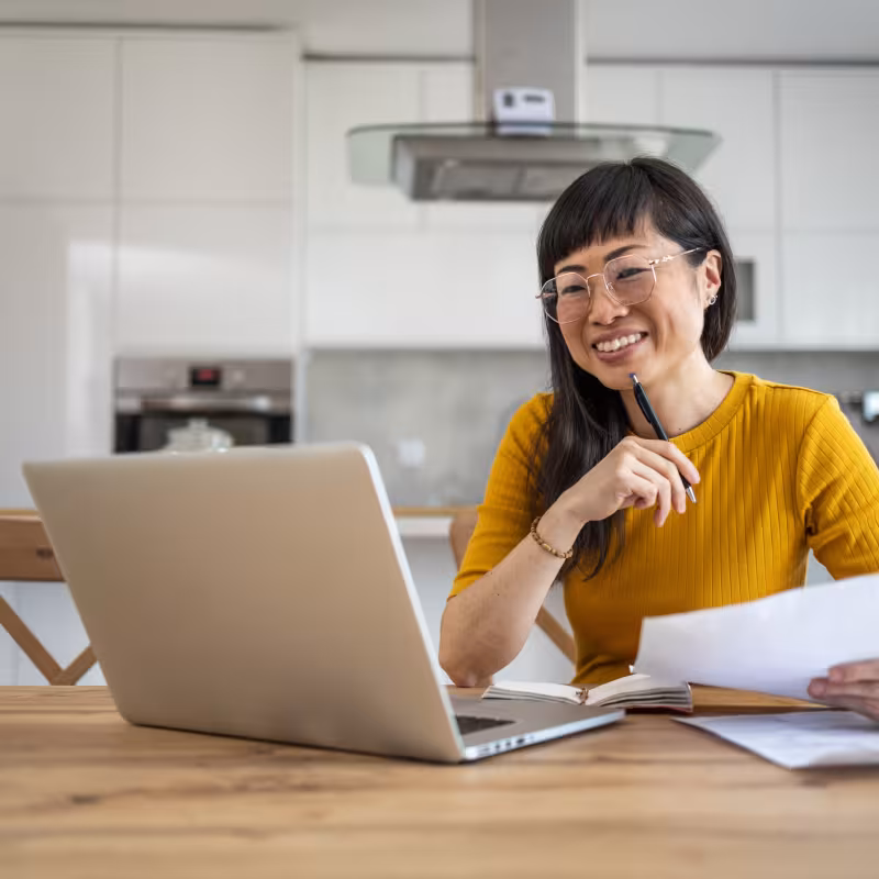 A woman sits at her dining table with her laptop and some papers out as she looks at something on the screen