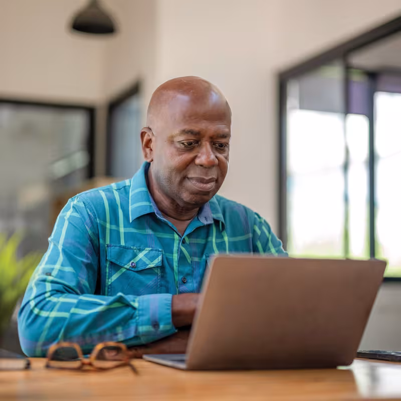 A senior man sits at a computer, his glasses on the desk next to him. 