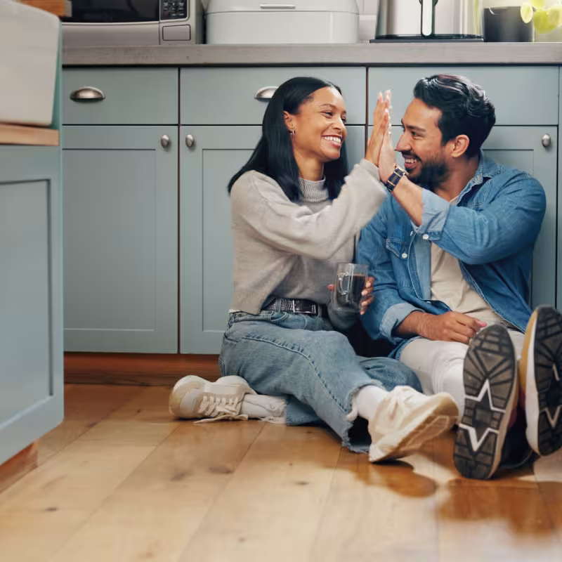 A young couple sits on the floor of their kitchen drinking coffee and sharing a high five 