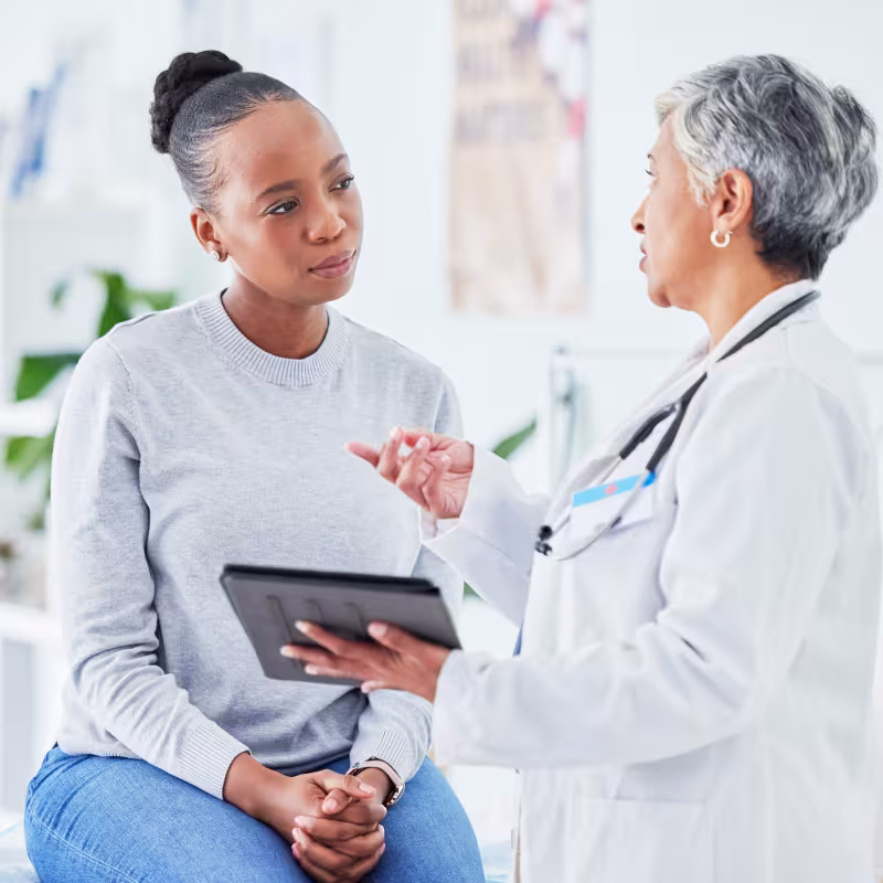 A woman sits on an exam table listening to a doctor who is speaking to her and holding a tablet in her hand. 