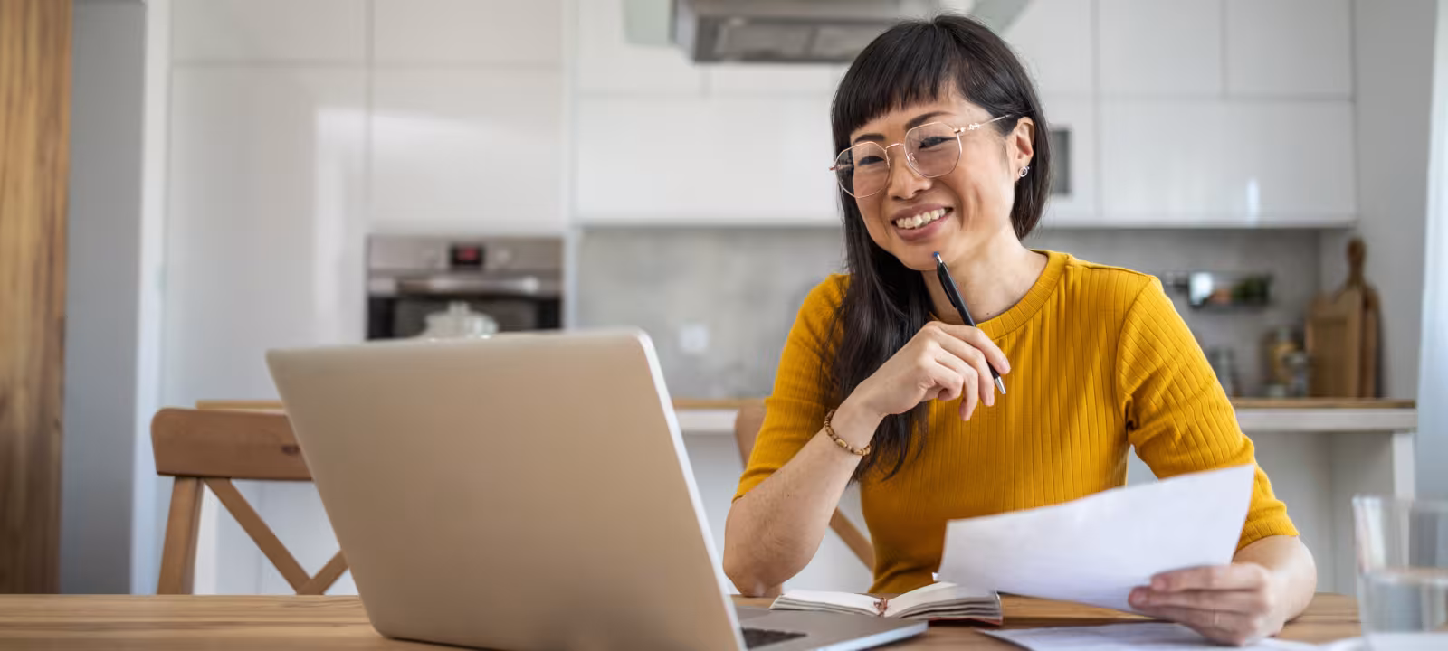 A woman sits at her dining table with her laptop and some papers out as she looks at something on the screen