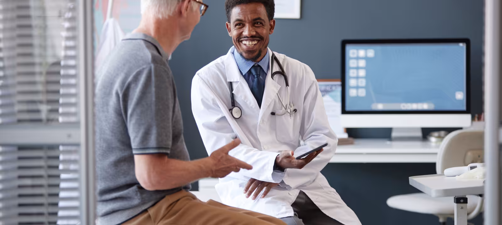 An older man sits on an exam table with his back to the viewer, a doctor in a lab coat sits casually facing the man and smiling. 