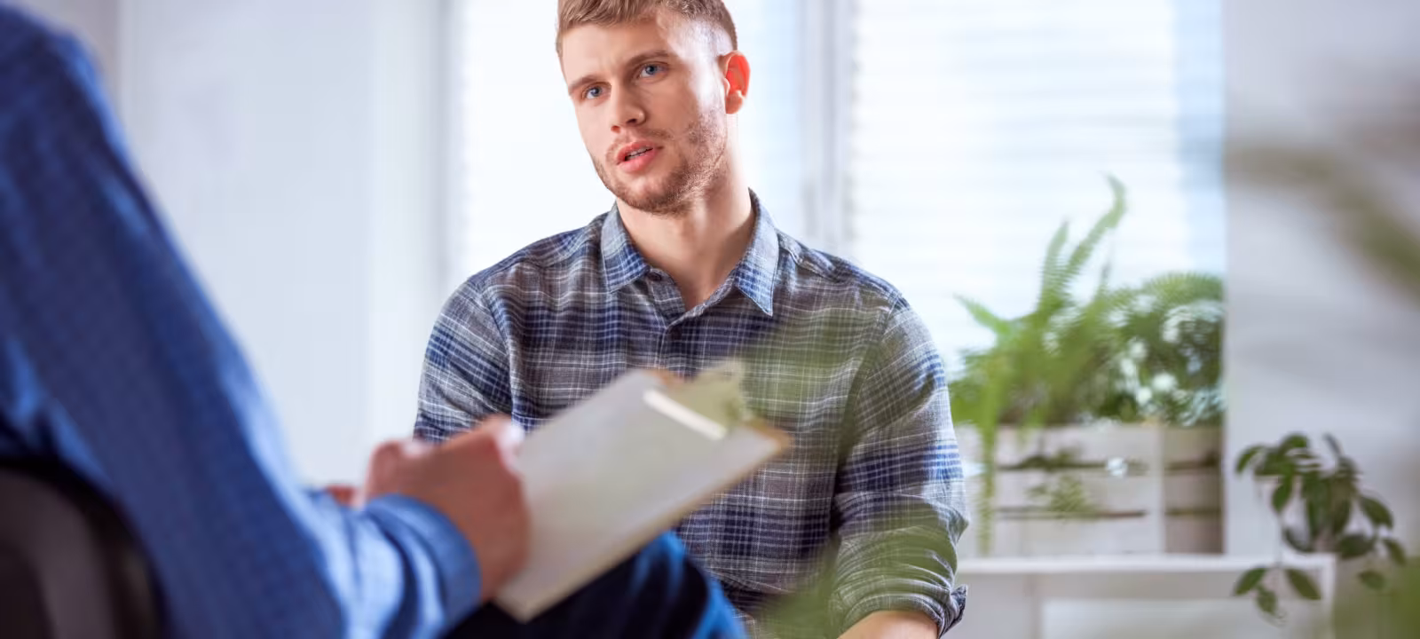 A young adult male sits in a therapist office, the therapist is out of frame but you can see their arm and a notepad for taking notes while the patient speaks.