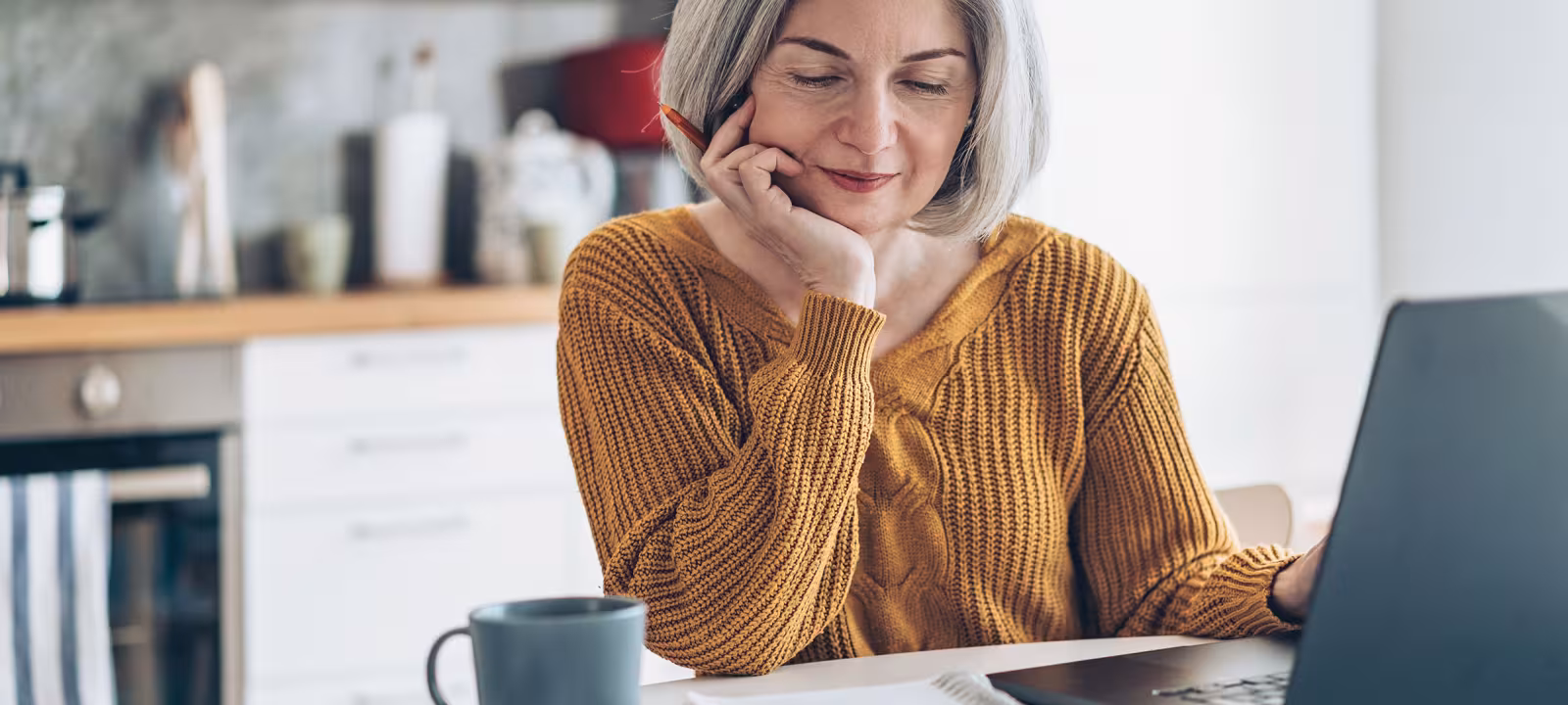 An older woman with grey hair in a golden sweater sits at a computer with a notebook, pen, and coffee mug looking contemplatively at the screen
