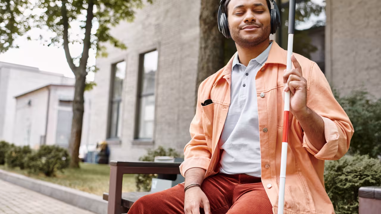 A cheerful young man with vision impairment enjoys music in a sunny outdoor setting with his cane. 