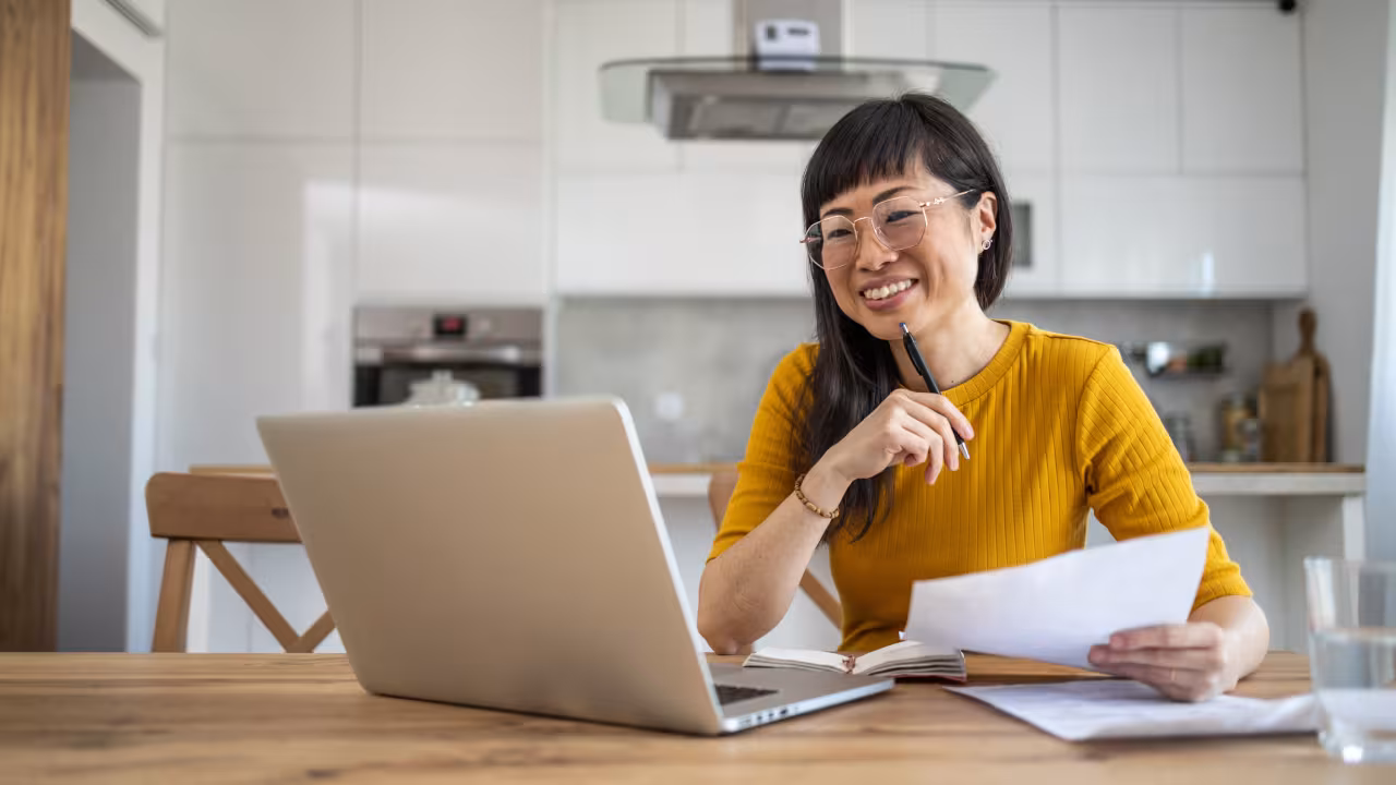A woman sits at her dining table with her laptop and some papers out as she looks at something on the screen