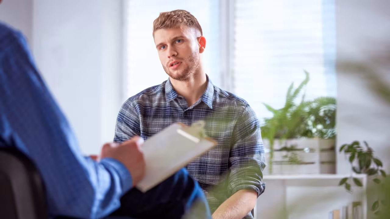 A young adult male sits in a therapist office, the therapist is out of frame but you can see their arm and a notepad for taking notes while the patient speaks.