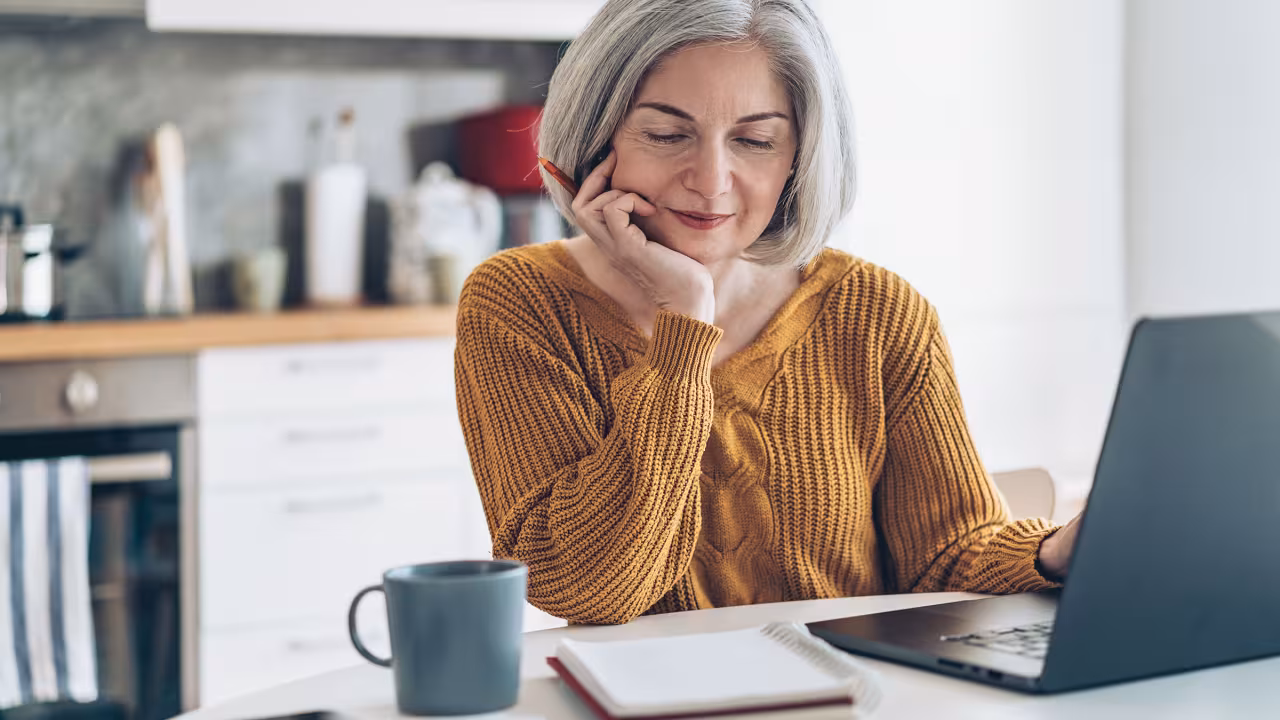An older woman with grey hair in a golden sweater sits at a computer with a notebook, pen, and coffee mug looking contemplatively at the screen